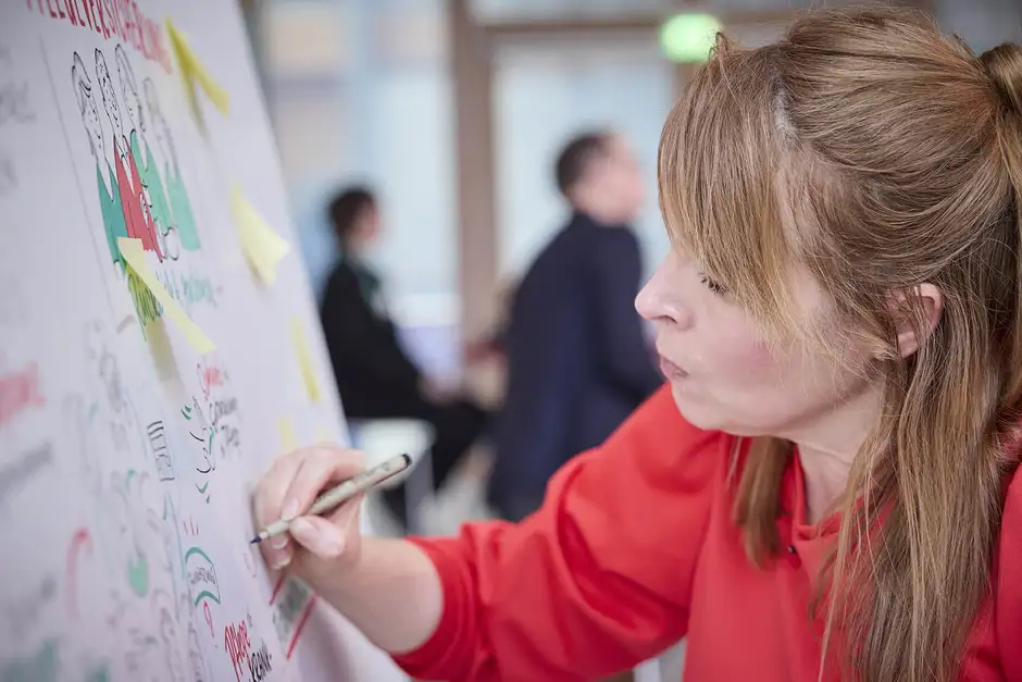 Graphic Recording Berlin &ndash; Nadine Ro&szlig;a in rotem Pullover zeichnet konzentriert an einem Graphic Recording f&uuml;r den Fachkongress '30 Jahre Pflegeversicherung'.