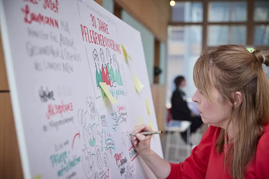 Graphic Recording Berlin &ndash; Nadine Ro&szlig;a in rotem Pullover zeichnet konzentriert an einem Graphic Recording f&uuml;r den Fachkongress '30 Jahre Pflegeversicherung'.
