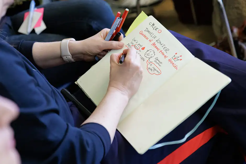 Person macht eine Sketchnote in ein aufgeschlagenes Notizbuch. Mit farbigen Stiften (rot, blau, schwarz) wird eine visuelle Mitschrift zu einem Vortrag &uuml;ber Menstruationsgesundheit erstellt. Auf der Seite steht unter anderem &bdquo;Queen of Menstrual Health&ldquo; sowie ein gezeichneter Kopf mit Sprechblase. Die Person tr&auml;gt eine Smartwatch und sitzt inmitten anderer Zuh&ouml;rer.
