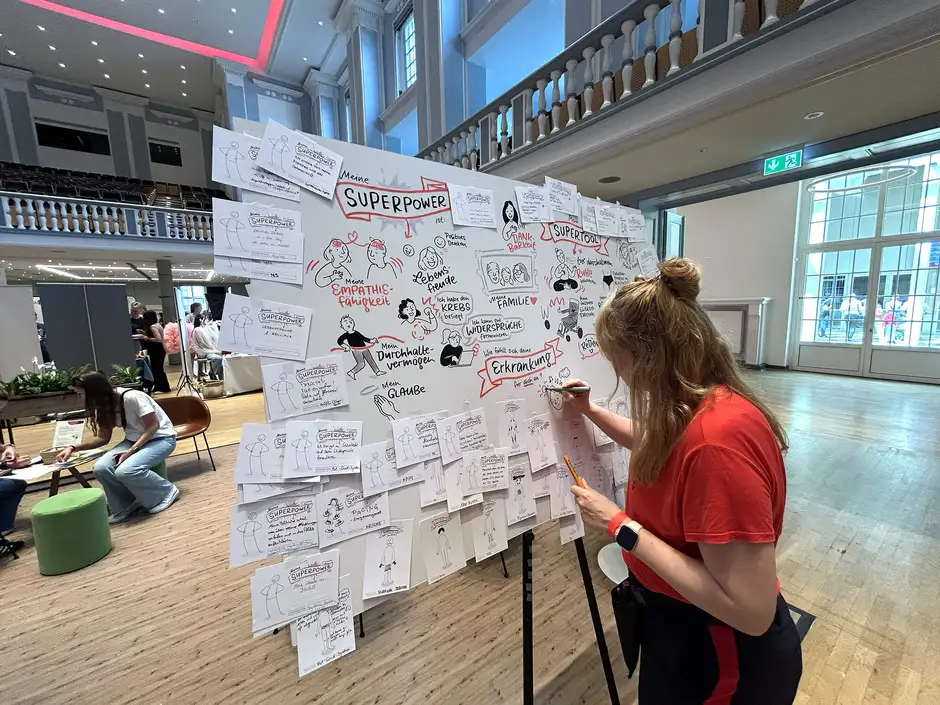 Live-Zeichnen beim K&auml;mpferherztreffen 2025: Nadine Ro&szlig;a erstellt Graphic Recording mit Sketchnotes an einer gro&szlig;en Pinnwand. Event-Zeichnen dokumentiert die Ideen und Superkr&auml;fte der Teilnehmer:innen.