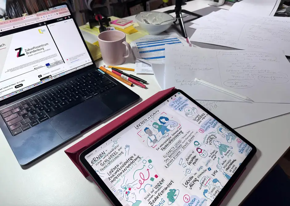 Schreibtisch mit Laptop, Tablet und Sketchnote zum Thema &bdquo;Lernen im Fokus&ldquo;, daneben Notizen, Stifte und Kaffeetasse w&auml;hrend eines Graphic-Recording-Prozesses